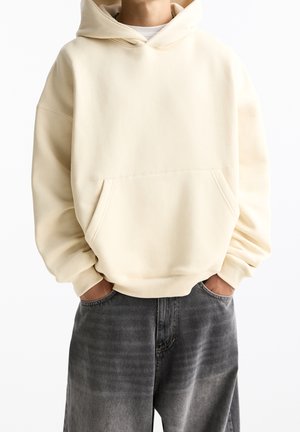 Felpa color crema con tasca a marsupio, tessuto morbido, vestibilità rilassata e polsini a coste, abbinata a jeans grigi scuri e sbiaditi.