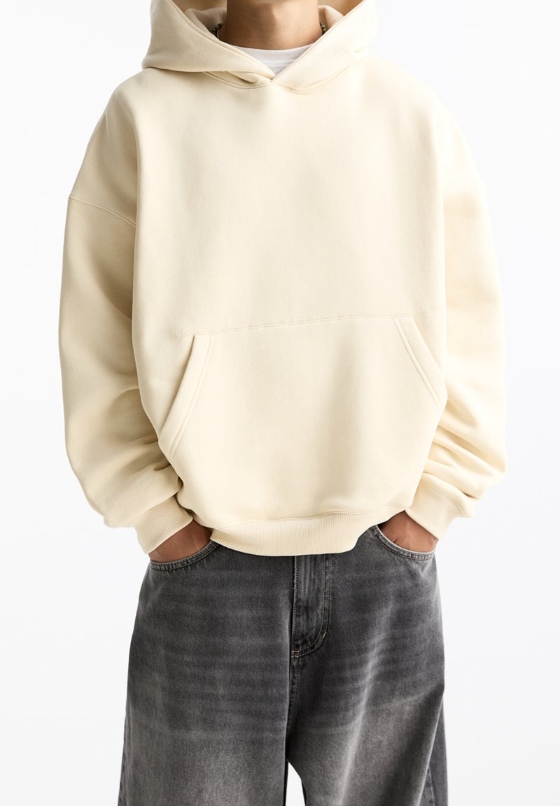 Krémová mikina s klokaní kapsou, měkká textura, volný střih a žebrované manžety, kombinovaná s tmavě šedými, vybledlými džíny.