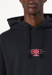 Hoodie marine avec un logo avant en rouge et des accents enflammés. Le matériau semble doux ; le design inclut des cordons de serrage et une poche kangourou.
