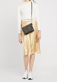 Mujer lleva una blusa blanca de manga larga, falda midi dorada brillante, zapatillas blancas y un bolso cruzado de piel de serpiente negra.