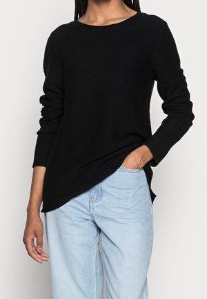 Person, die einen schwarzen Pullover mit langen Ärmeln und Bootsausschnitt sowie hellblaue Jeans trägt, eine Hand in der Tasche, vor weißem Hintergrund stehend.