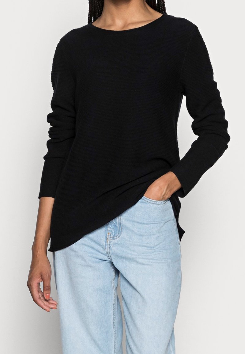 Personne portant un pull noir à manches longues avec un col bateau et un jean bleu clair, une main dans la poche, debout devant un fond blanc.