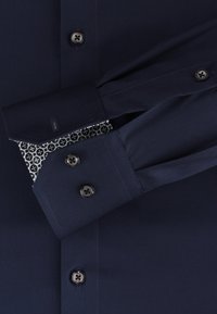 Navyblaues Hemd mit glatter Textur, schwarzen Knöpfen und gepolsterten Manschetten im weißen und dunkelblauen geometrischen Design.