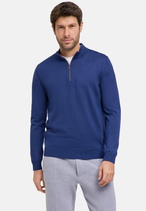 Blauwe zip-vrouw trui in een zachte brei textuur, met een korte rits en aangesloten manchetten. Gecombineerd met lichtgrijze broek.