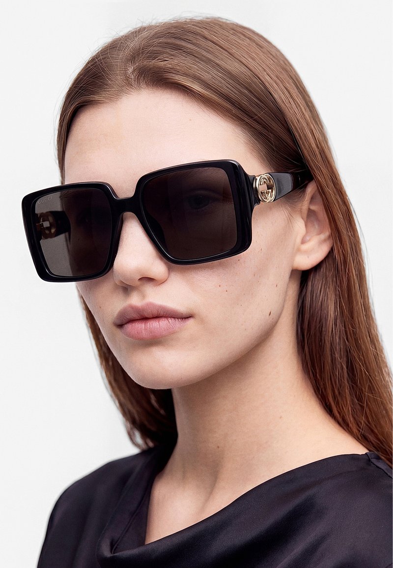 Junge Frau mit glattem braunem Haar trägt große schwarze quadratische Sonnenbrille und ein schwarzes seidiges Top vor hellem grauem Hintergrund.