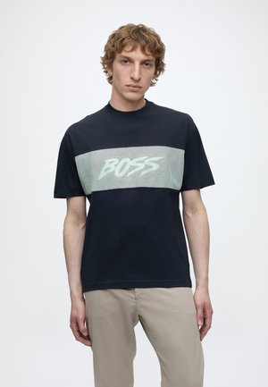 Νέος άνδρας με σγουρά μαλλιά φοράει μαύρο T-shirt με λευκή οριζόντια ραμμένη ταινία και το κείμενο "BOSS", συνδυασμένο με μπεζ παντελόνια σε απλό φόντο.