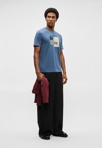 T-shirt de algodão azul com um padrão geométrico em creme e azul marinho. Calças largas pretas e um moletom bordeaux segurado à mão. Sapatos pretos.