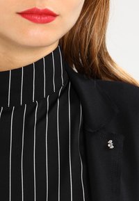 Czarna bluzka w pionowe białe paski, z wysokim kołnierzem, wykonana z miękkiego materiału, w połączeniu z czarnym żakietem. Srebrny pin w kształcie kwiatu jako akcent na żakiecie.