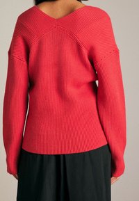 Czerwony prążkowany sweter dziany z dekoltem z tyłu w kształcie litery V i szerokimi rękawami, o teksturowanej powierzchni i kontrastującym czarnym dole.