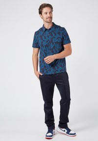 Marinblå polo med blå bladmönster, korta ärmar och knappkrage. Kombinerad med mörka byxor och blå, vita och röda sneakers.