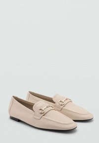 Beige loafers dengan tekstur halus, desain ujung runcing, sol datar, dan aksen perangkat keras logam bernuansa emas di bagian atas.