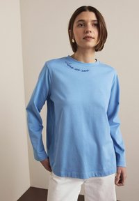 Modra majica z dolgimi rokavi in okrolim izrezom, na kateri je temno modro izvezena napis "Izgubljen na morju". Mehka tekstura, sproščen kroj, kombinirana z belimi hlačami.