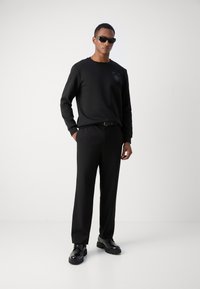 Sweatshirt noir avec un accent texturé subtil, associé à un pantalon noir ample et des chaussures noires, complété par des lunettes de soleil foncées.