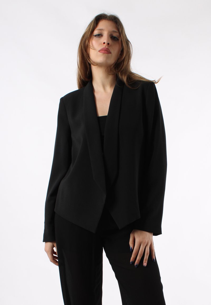 Blazer nero realizzato in un tessuto liscio, con un design frontale drappeggiato, maniche lunghe e una vestibilità sartoriale. Indossato sopra un top nero.