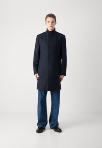 Cappotto in misto lana colore blu navy con colletto alto, chiusura a cinque bottoni, tasche frontali e silhouette dritta. Abbinato a pantaloni in denim a gamba larga.