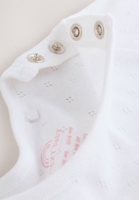 Vêtement blanc pour bébé avec un petit motif perforé et trois boutons-pression métalliques partiellement ouverts sur un tissu doux.