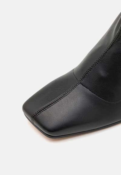 Botină din piele neagră, cu vârf pătrat, textură netedă și detaliu de cusătură subtil pe margine. Proiectată pentru o potrivire elegantă.