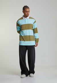 Svītrainā garo piedurkņu polo krekls gaiši zils un olīvu zaļš ar baltu apkakli, kombinēts ar platām melnām biksēm un baltām kedām.