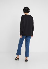 Person with short black hair wearing a black long-sleeve shirt, blue cropped jeans with frayed hems, and black heeled mules, facing away.