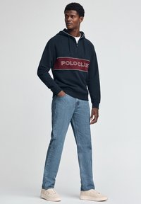 Sudadera con capucha azul marino con una franja horizontal burdeos con el logo "POLOCLUE", combinada con jeans azul claro y zapatillas beige.