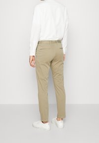 Person klädd i beige chinos, vit skjorta och vita sneakers, stående vänd mot en enkel ljusgrå vägg.