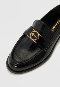 Schwarze Lackleder-Loafers mit glatter Textur, die ein goldenes Emblem am Riemen aufweisen. Rundes Fußteil und flacher, niedriger Absatz.