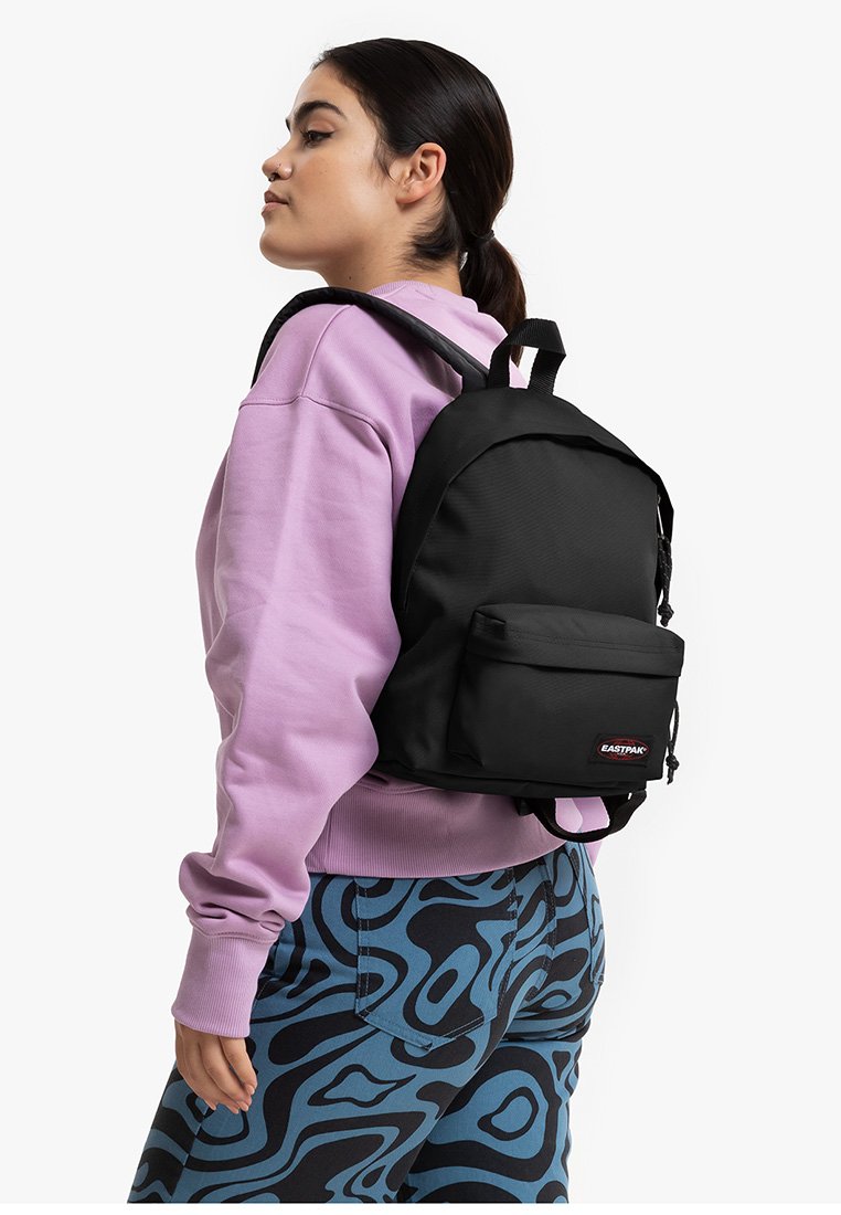 Zwarte rugzak met een gestructureerd ontwerp, voorzak met rits en verstelbare banden. Draagt door een persoon in een paarse sweatshirt en met een patroonbroek.