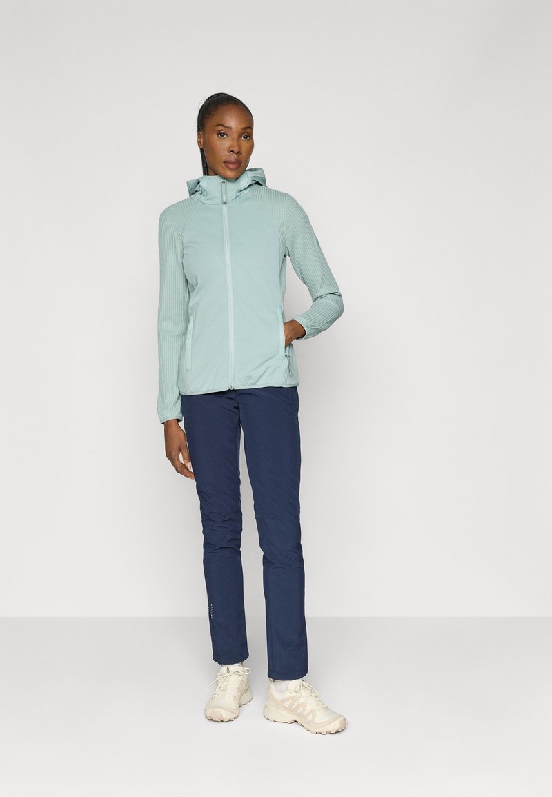 Helle blaue Zip-Jacke mit Kapuze und gerippter Textur, kombiniert mit dunkelblauen Hosen und hellfarbigen Schuhen, vor einem einfarbigen Hintergrund stehend.