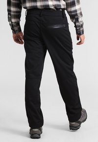 Czarne spodnie outdoorowe z płaskim pasem i kieszeniami na zamek, wykonane z gładkiego, lekkiego materiału. Noszone z szarymi butami do wędrówek.