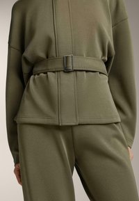 Set oblečenia v olivovej zelenej farbe obsahujúci štruktúrovaný top s opaskom v páse a kontrastným prešívaním, spárovaný s voľnými nohavicami.