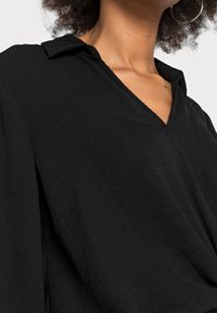 Schwarze, strukturierte Bluse mit tiefem V-Ausschnitt und Kragen. Mit lockeren Ärmeln und einem fließenden Schnitt, der ein lässiges Design betont.