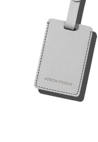 Étiquette de bagage en cuir gris avec des coins arrondis, des coutures visibles et le logo "HORIZN STUDIOS" en relief. Dispose d'une boucle de fixation sécurisée.