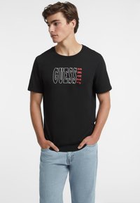 Juoda medvilninė marškinėliai su "GUESS JEANS" tekstu baltos ir raudonos spalvos. Trumpomis rankovėmis ir apvaliu iškirpimu. Derinami su šviesiai mėlynomis džinsinėmis kelnėmis.