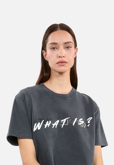 T-shirt en coton gris avec un col rond et des manches courtes, ornée d'un grand texte graphique blanc "QU'EST-CE QUE C'EST ?" sur le devant.