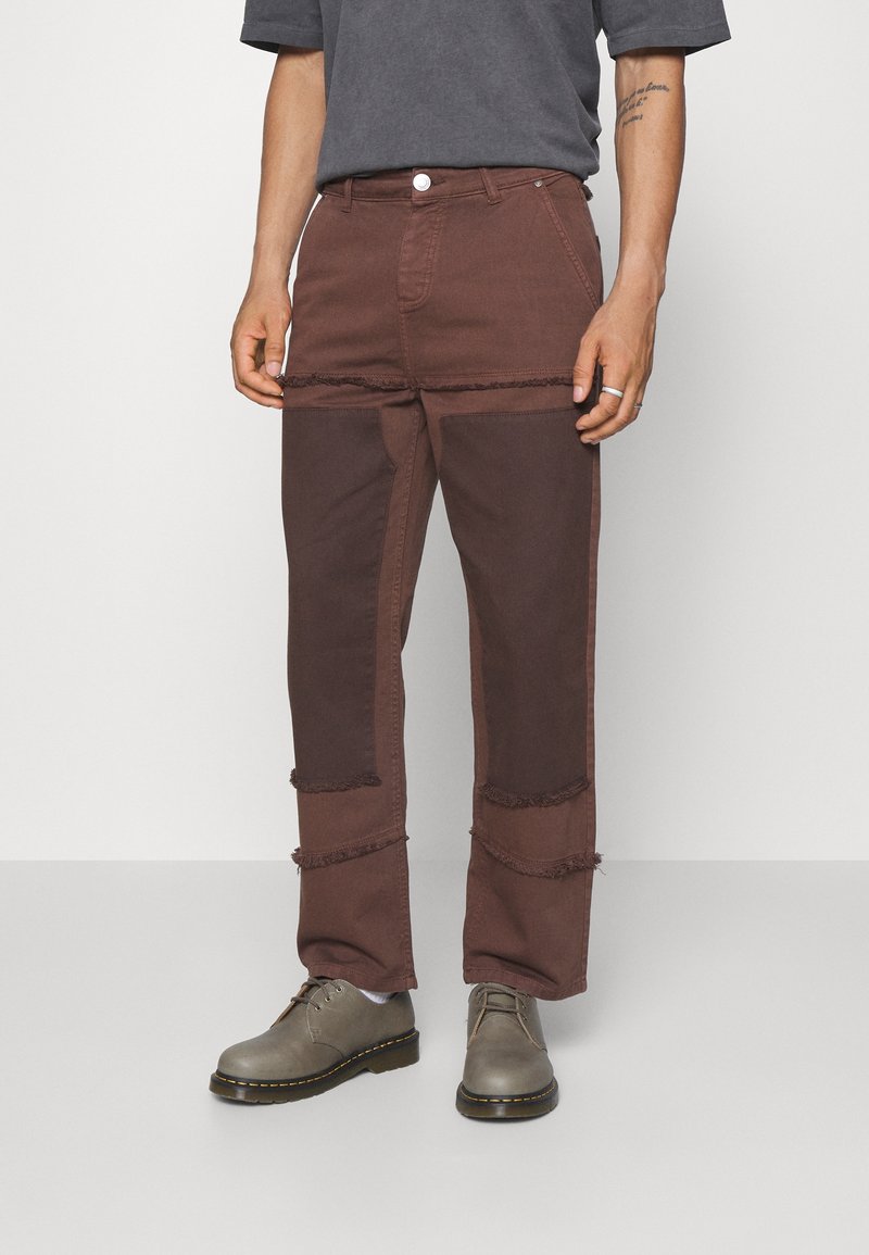 Pantalon en denim marron présentant un mélange de textures avec des accents effilochés. Conçu avec une coupe ample et associé à des chaussures grises à lacets.