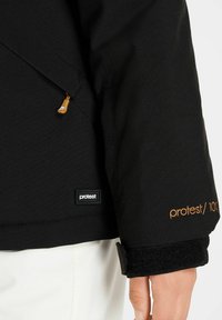 Giacca esterna nera con texture liscia, dotata di tasca con zip e polsini in velcro. Logo "Protest/10K" in oro sulla manica.