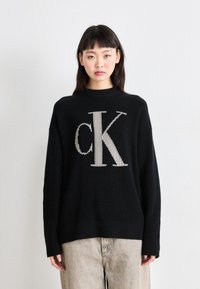 Črn pleteni pulover s velikim bež "CK" logotipom na sredini, z rebrastim ovratnikom in manšetami. Ujemajoče se svetlo bež kavbojke.