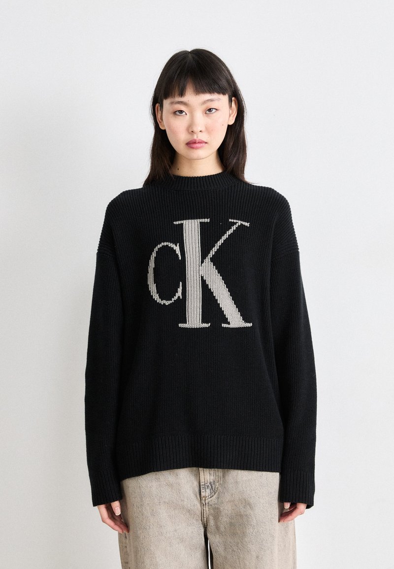 Črn pleteni pulover s velikim bež "CK" logotipom na sredini, z rebrastim ovratnikom in manšetami. Ujemajoče se svetlo bež kavbojke.