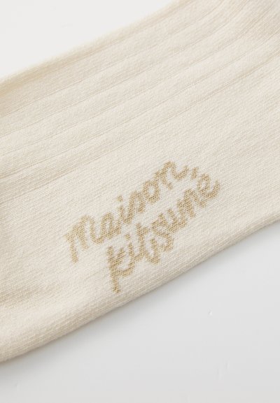 Zābaku augstuma krēma zeķe, kas izgatavota no maiga auduma, ar smalku ribotu struktūru un zelta izšūtu logotipu "Maison Kitsuné."