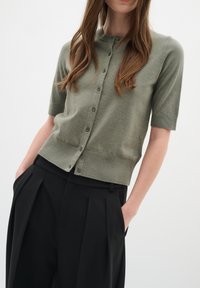 Cardigan a maniche corte di colore verde oliva, realizzato in materiale knit, con chiusura a bottoni e un design aderente. Abbinato a pantaloni neri a vita alta.