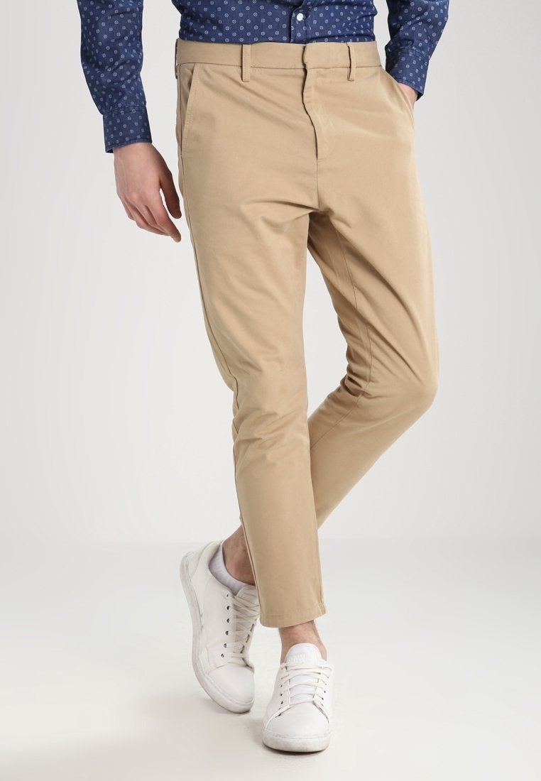 Beige pants with a slim fit, made of cotton fabric, featuring front pockets, and styled with white sneakers. The shirt has a patterned design.