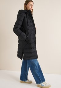 Kvinde iført en sort hætte-pufferjakke, blå vide jeans og hvide sneakers med blå detaljer, stående foran en ensfarvet baggrund.