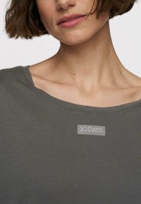Szara, lekkia bluzka z szerokim dekoltem i krótkimi rękawami. Posiada małą, wytłoczoną metkę z logo "10 DAYS" z przodu.
