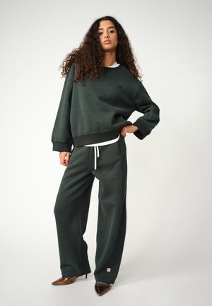Sweatshirt supradimensionat verde închis și pantaloni jogger dintr-un material moale, cu un maieu alb pe dedesubt și tocuri ascuțite maro ca încălțăminte.