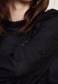 Schwarze Bluse mit detaillierter floralem Stickerei, Spitzenakzenten und einem Rüschenausschnitt. Lange Ärmel mit gerafften Bündchen. Glatte Textur.