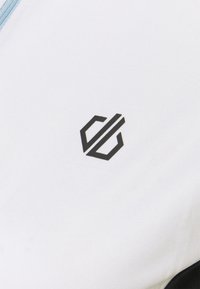 Hvidt stof med en tekstureret overflade præsenterer et sort geometrisk logo. Designet inkluderer en lyseblå lynlås ved kraven.