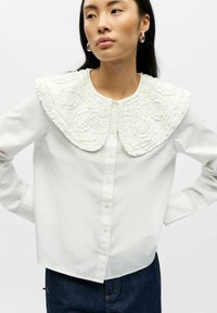 Blusa blanca de algodón con un gran cuello texturizado y con volantes que presenta patrones intrincados. Diseño con botones en la parte delantera y mangas largas.