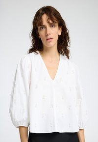 Blouse blanche à col en V avec des manches bouffantes, ornée d'une broderie florale texturée sur l'ensemble. Tissu léger avec une coupe décontractée.
