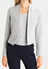 Woman wearing light gray open-front blazer over a light gray patterned shirt and dark navy pants, standing against a white background.
