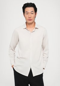 Lys beige langermet skjorte med krage som kan kneppes, med en glatt tekstur og en svak glans. Diskret logo brodert på brystet.